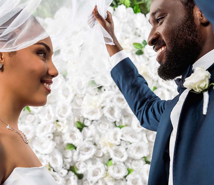 View of happy african american bridegroom touching white veil and smiling near bride and flowers.