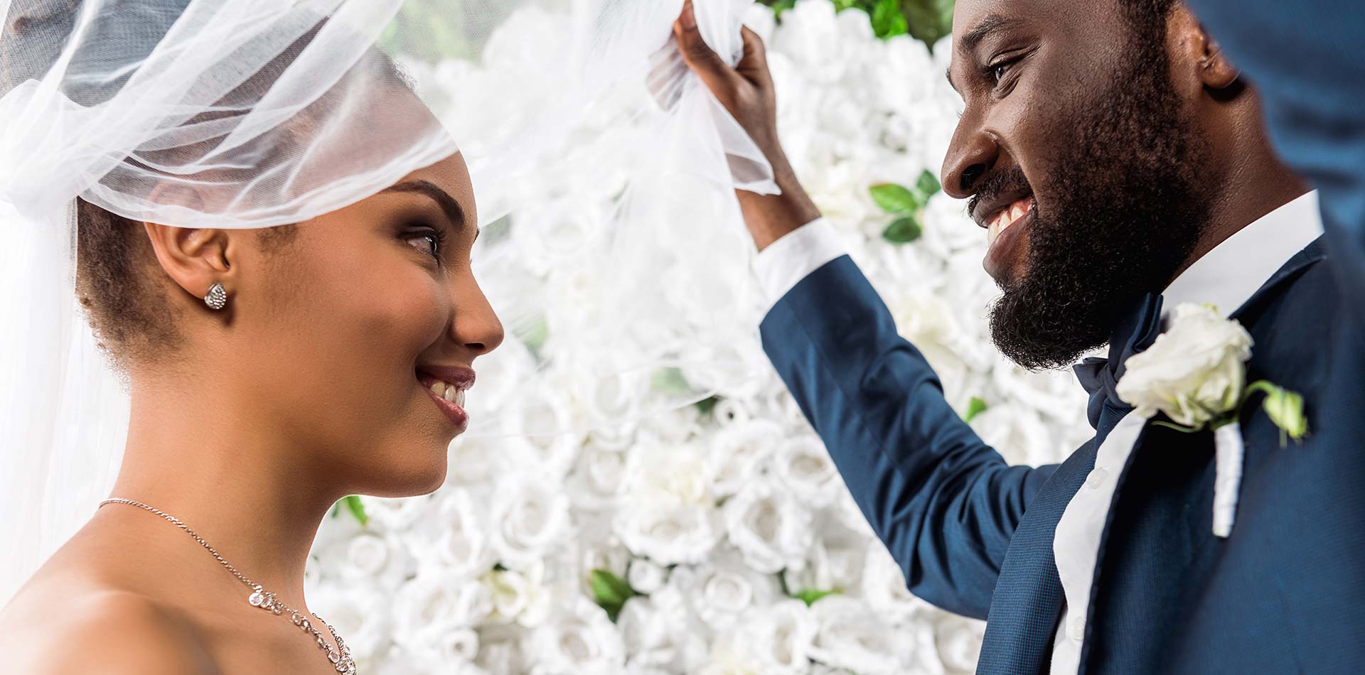 View of happy african american bridegroom touching white veil and smiling near bride and flowers.