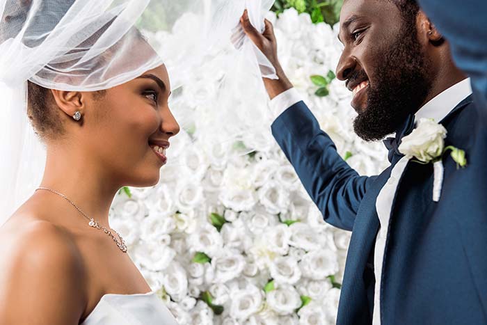 View of happy african american bridegroom touching white veil and smiling near bride and flowers.